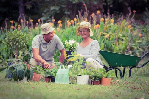 A mature couple in a lush garden, sitting on the grass and engaging in gardening activities. The man, wearing a light grey T-shirt and a wide-brimmed hat, is smiling and leaning toward the woman, who is dressed in a light grey top and a straw hat, also smiling. They are surrounded by various potted plants of different sizes and types, including flowering plants and leafy shrubs, placed on the neatly maintained lawn. Behind them, there is a flower bed filled with blooming purple, orange, and yellow flowers, creating vibrant colour contrasts against the green foliage. To the right, a sheaf of gardening tools, such as a watering can and a small spade, are visible, indicating their gardening work. The scene is set in an outdoor garden with natural light, suggesting a bright, possibly sunny day suited for outdoor maintenance and gardening services in Sidcup, as part of a well-kept landscaped yard accessible via postcode related to Sidcup. The overall environment reflects a peaceful and productive gardening space supported by local gardening experts like Hedge Trimming Sidcup.
