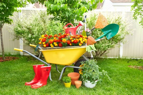 A colourful backyard garden scene featuring a yellow wheelbarrow filled with vibrant red and yellow flowering plants, positioned on a well-maintained green lawn. Surrounding the wheelbarrow are gardening tools, including a red watering can, a green trowel, and an orange spade, with additional planting containers such as small terracotta pots and a white bucket with a leafy plant. In the background, a wooden fence and lush trees provide privacy and natural shade. The garden appears to be in a suburban residential area, typical of Sidcup, with a neat, organized layout suitable for gardening and outdoor maintenance services offered by Hedge Trimming Sidcup. The scene suggests an environment ready for gardening activities, with a balance of flowered beds, grass, and garden equipment, under natural daylight with clear weather conditions typical of spring or summer in the UK.