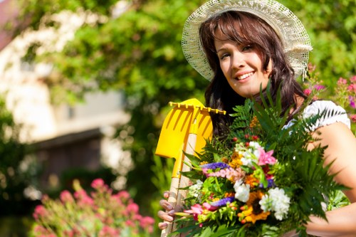 A woman with shoulder-length brunette hair wearing a wide-brimmed straw hat is smiling and standing in a well-maintained garden during daylight. She is holding a vibrant bouquet of flowers with various colours, including pink, white, purple, and yellow, along with green foliage, suggesting recent gardening or planting activity. Behind her, the garden features a lush, green lawn with dense trees and shrubbery, creating a natural backdrop. The lawn appears neatly mown with textured grass exhibiting different shades of green. To her right, there is a flower bed edged with soil and partially visible pink blossoms, indicating a well-organised garden layout. The environment suggests a typical outdoor space in Sidcup or a similar area, with sunlight filtering through the trees, casting soft light and gentle shadows across the scene. The overall setting reflects a tidy, cared-for garden that benefitted from professional gardening services, such as hedge trimming or lawn maintenance, indicative of the offerings at hedgetrimmingsidcup.co.uk.