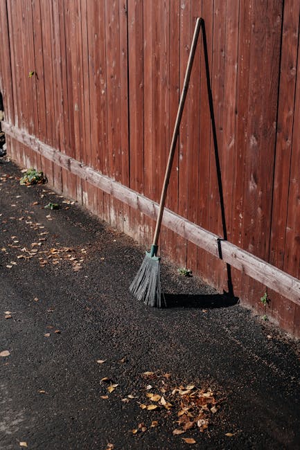 A broom with a long wooden handle and grey bristles, leaning against a red wooden fence in a garden setting. The ground is covered with black asphalt and scattered dry leaves. The fence is composed of